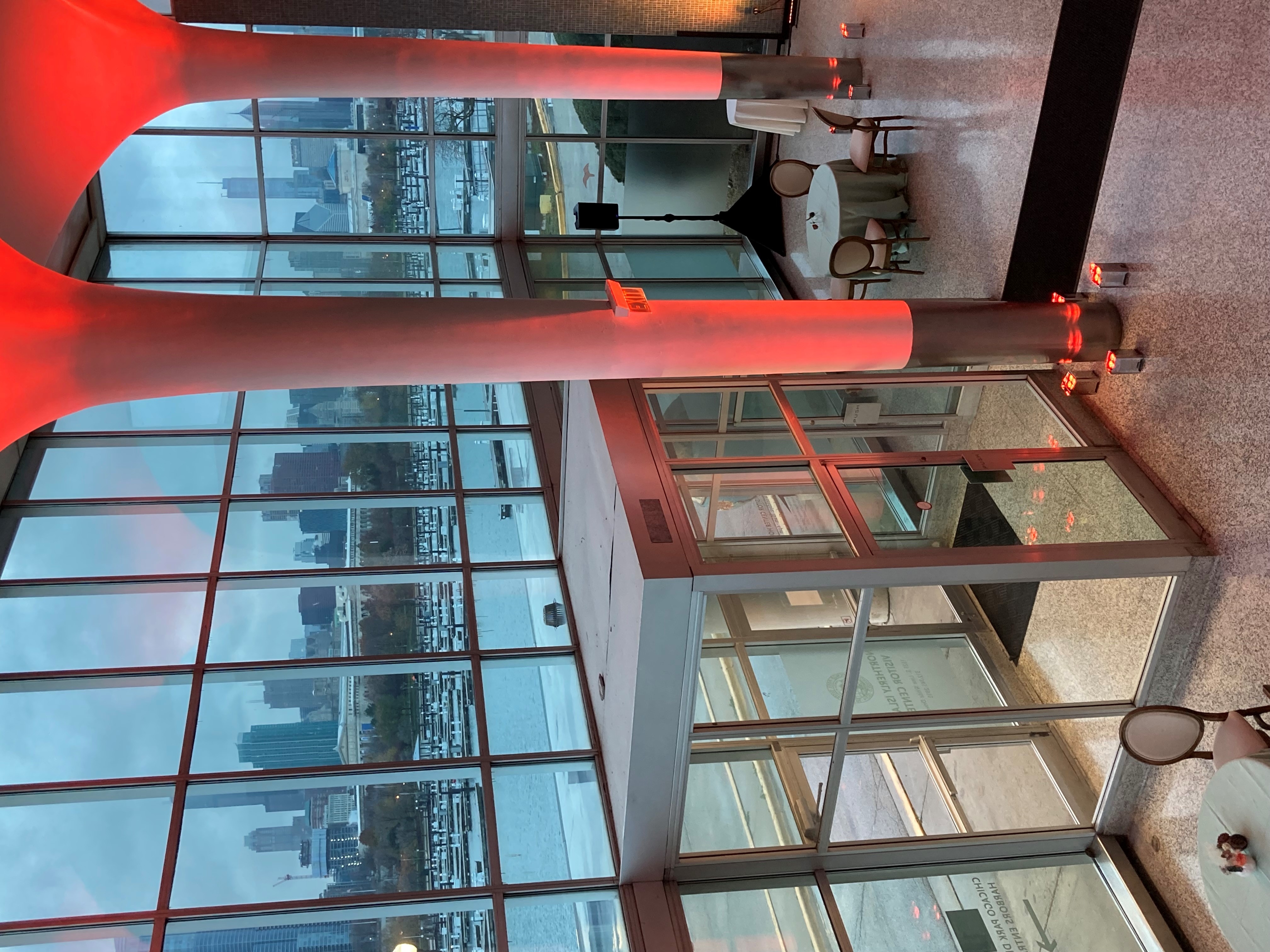 Red-lit columns beside large windows overlooking a cityscape and harbor. Small cafe table in foreground.